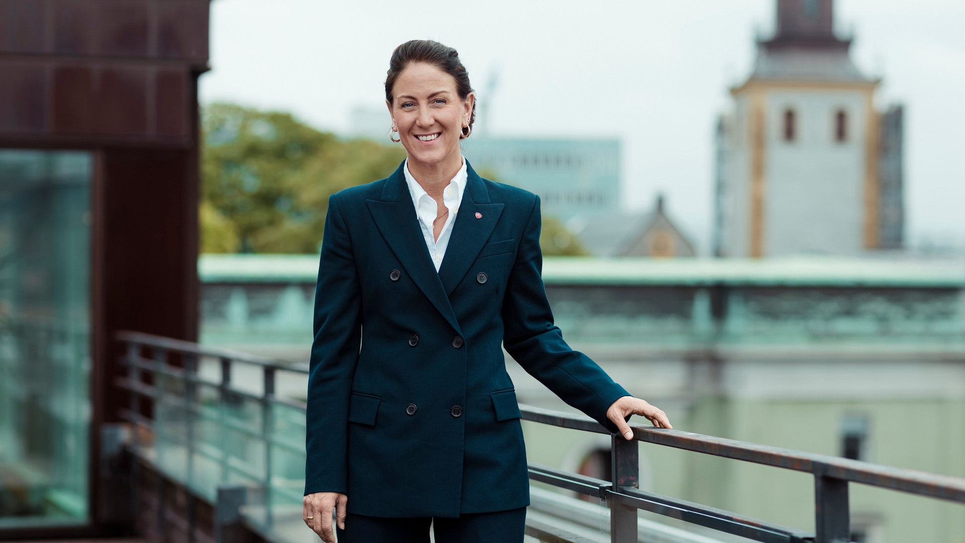 Arbeidsministeren Tonje Brenna står i sort dress på en veranda, du kan skimte andre bygninger i bakgrunnen