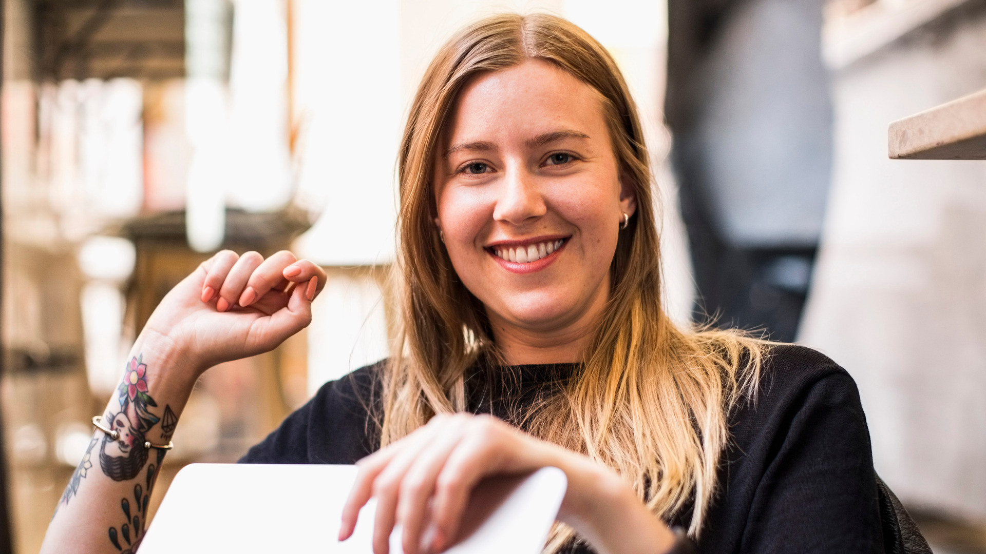 Ung jente sitter med en PC i fanget, tatoveringer på begge armene og jobber i et åpent landskap