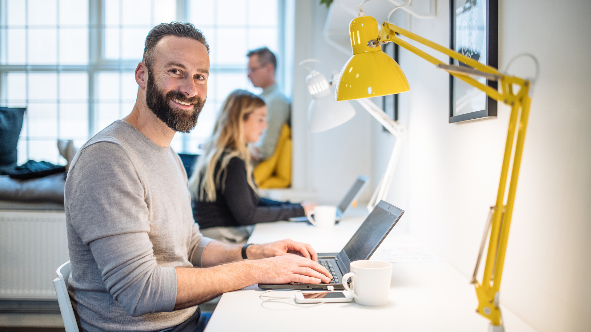 Mann sitter ved skrivebord med en gul lampe og skriver på PC.