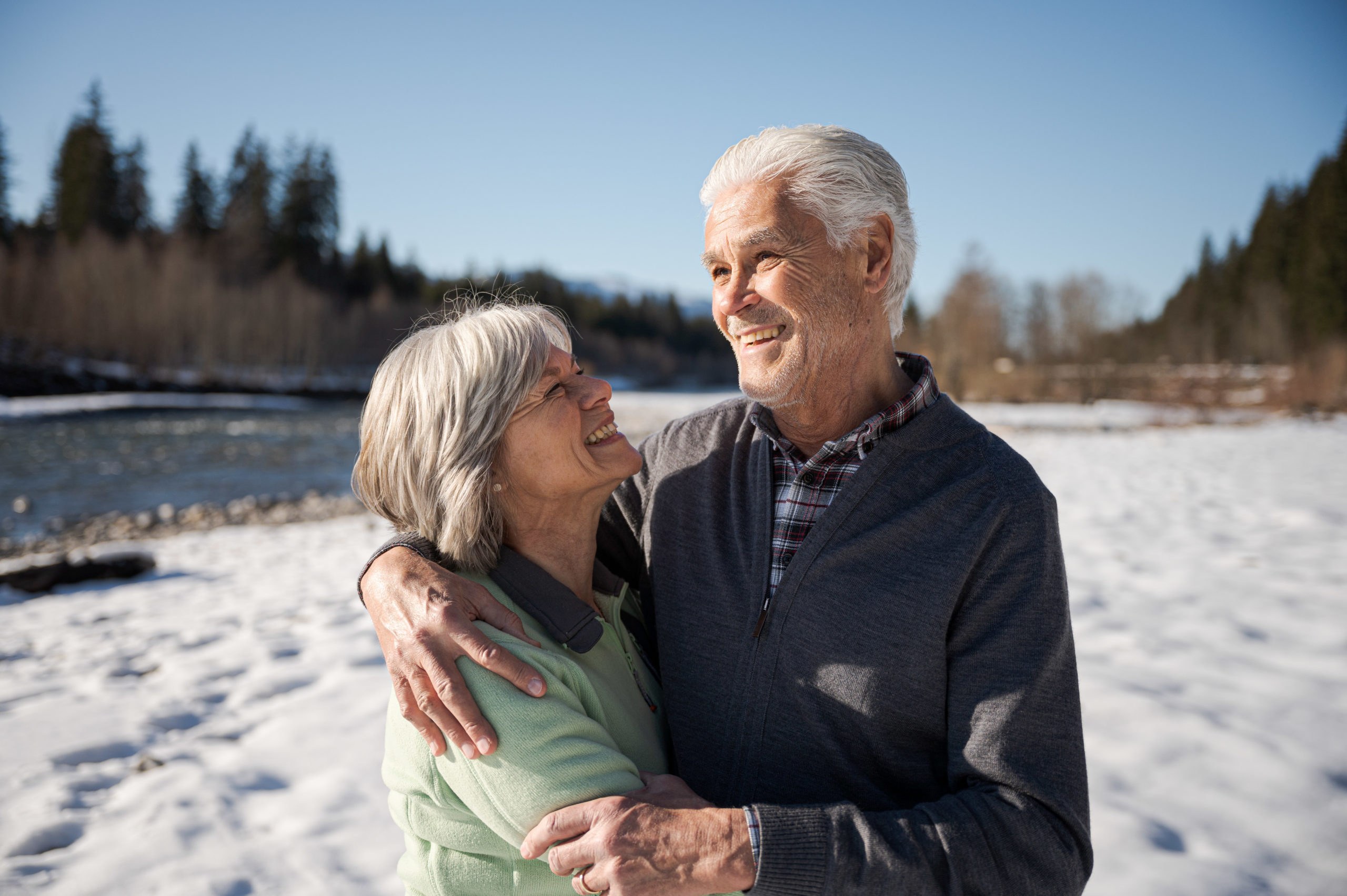 Eldre par holder rundt hverandre ved vannet med snø på bakken.