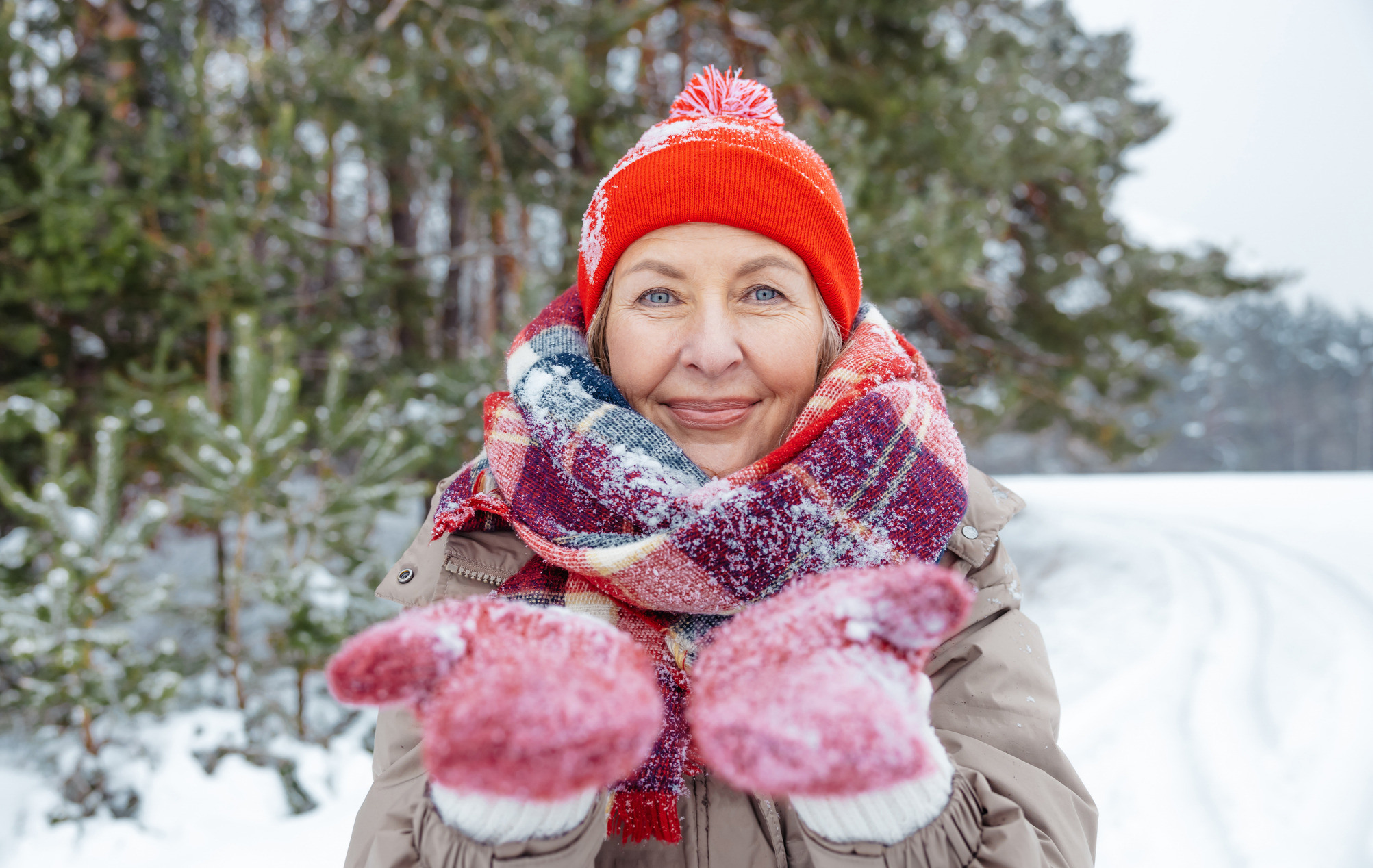 Kvinne i rød lue og røde votter viser frem snøen på vottene og ser mot kamera.