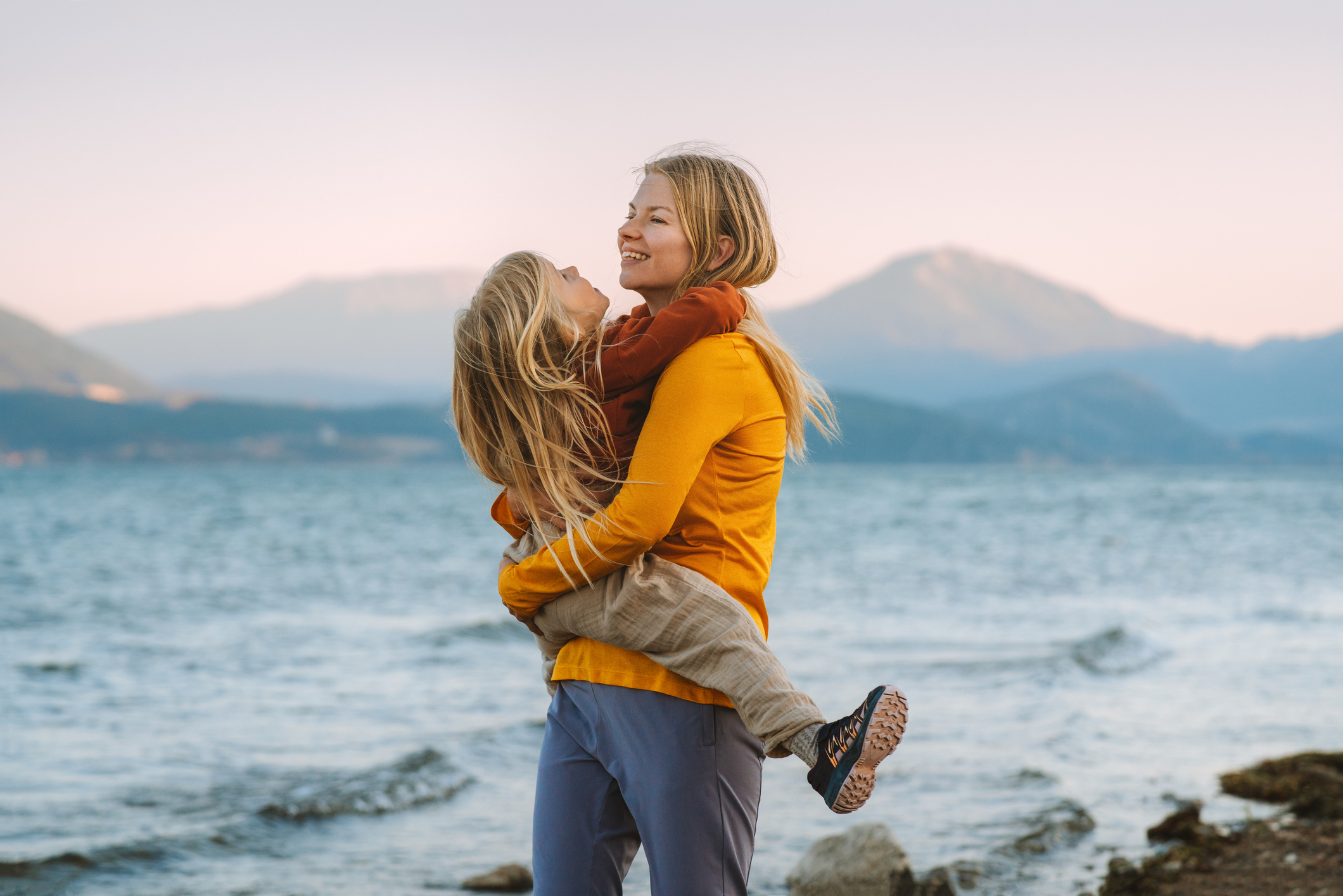 Ung mor holder datter ca 4 år i armene foran en strand med fjell i bakgrunnen